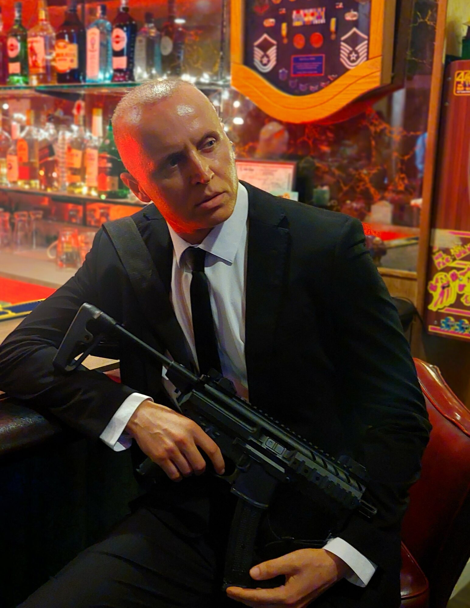Man in suit holding a firearm, sitting in a colorful bar or arcade setting.