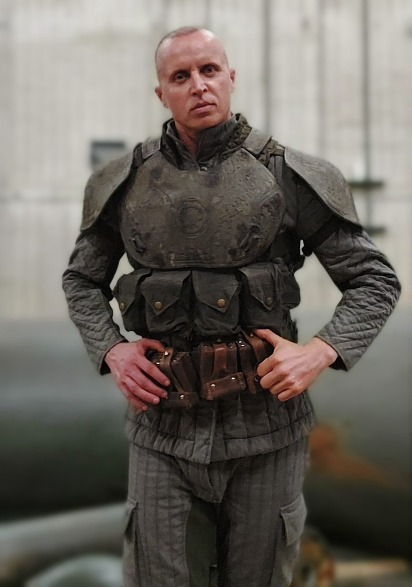 Person in military-style uniform with armor, standing outdoors with hands on hips, serious expression, gray background.