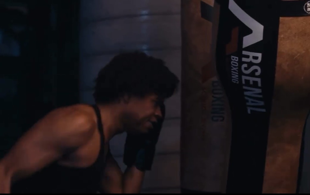 Person with curly hair in a gym, lifting a punching bag, wearing a sleeveless top.