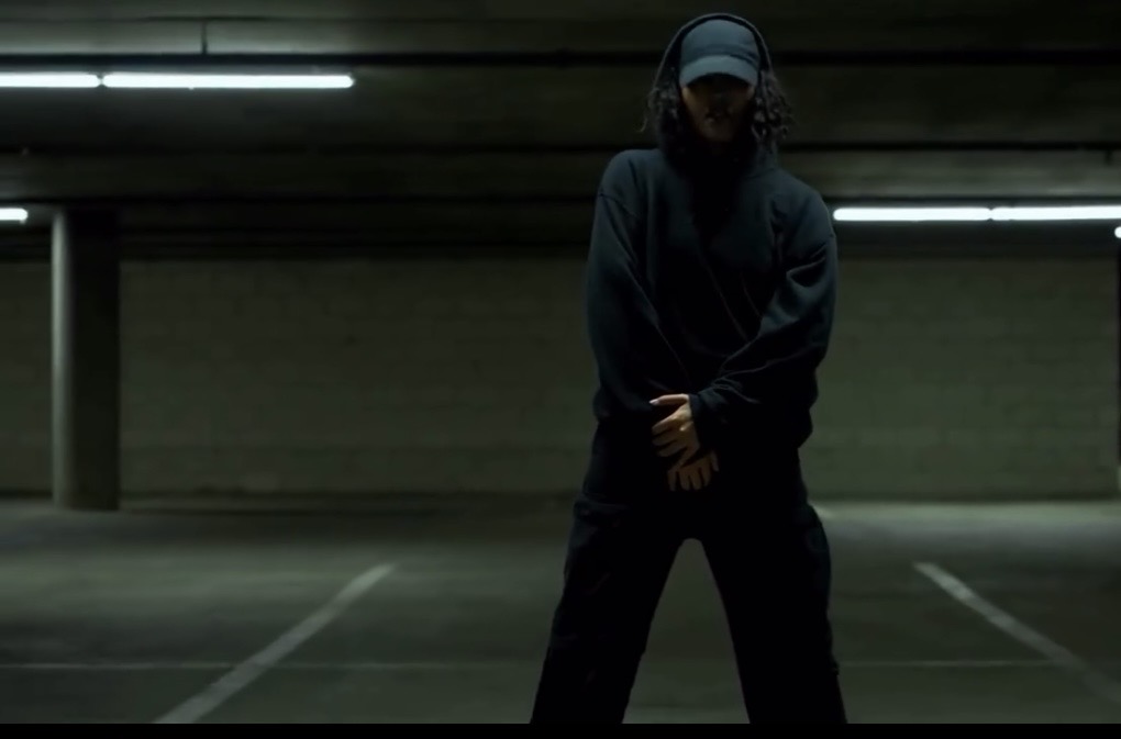 Person standing in a parking garage wearing dark clothing and a cap, with hands clasped in front.