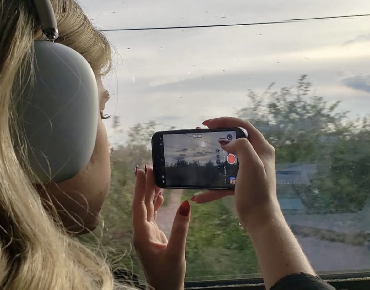 Person with headphones taking a photo with a smartphone on a bus or train, window showing trees and sky.