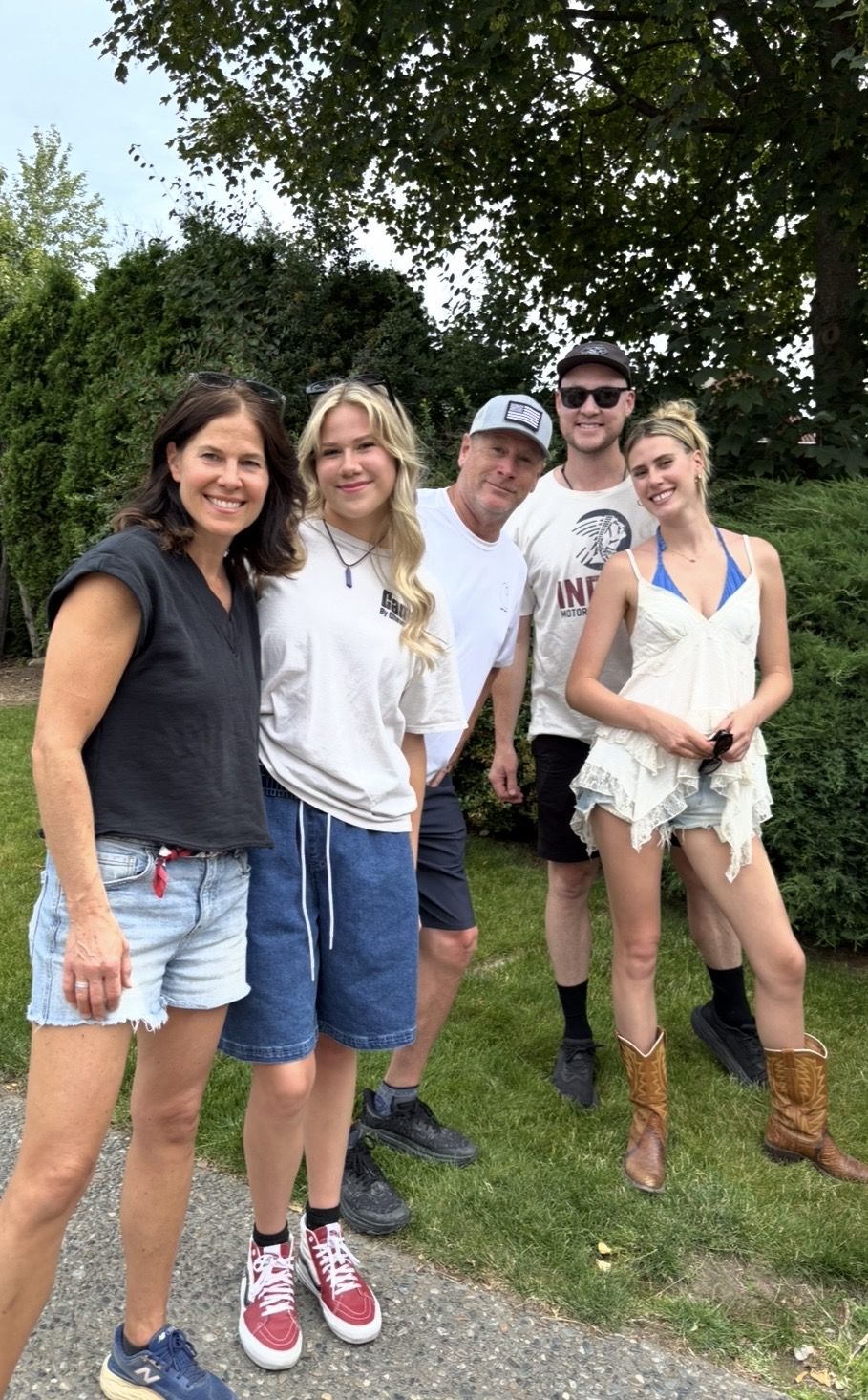 Five people standing outdoors on grass, smiling, with trees and bushes in background.