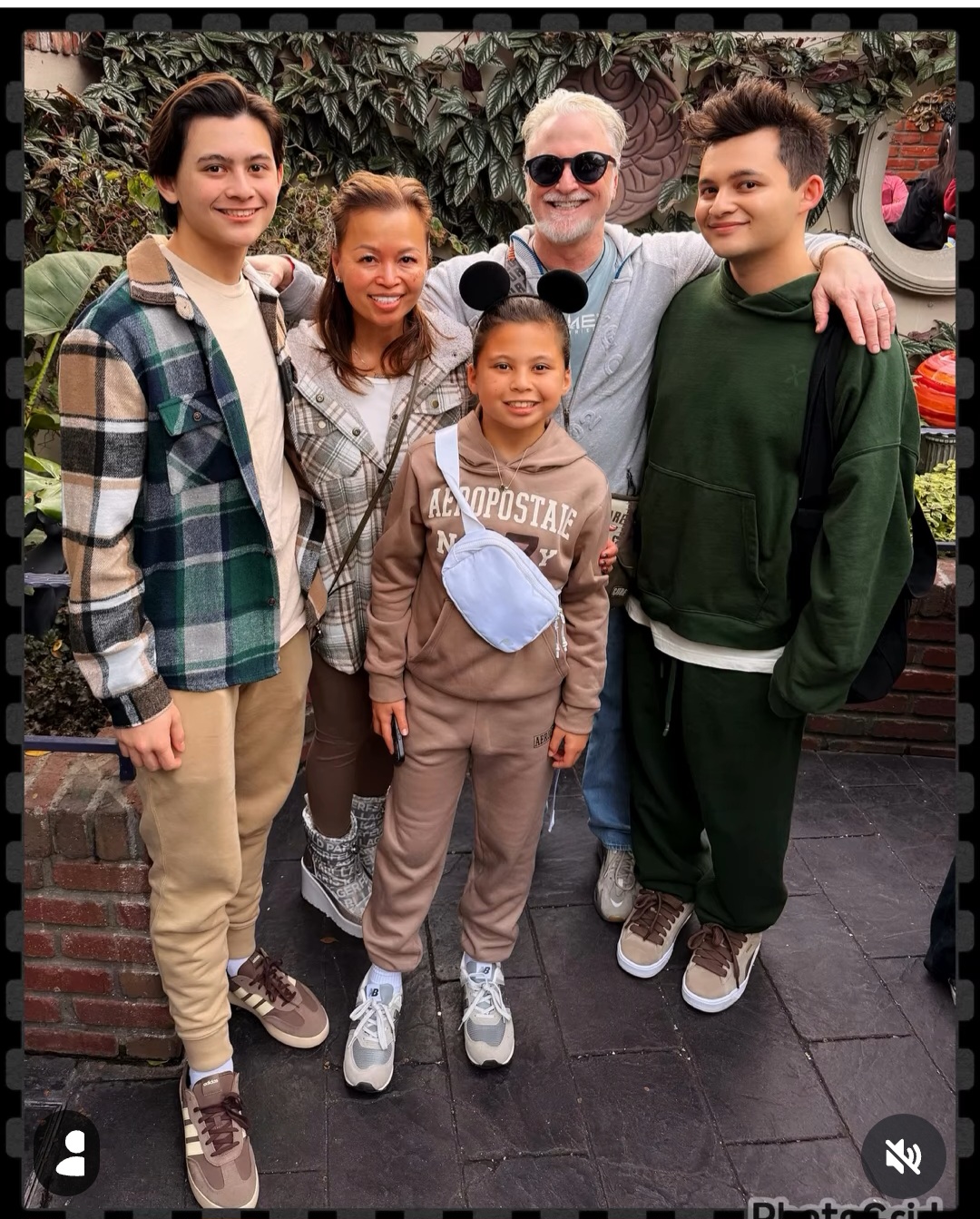 Group of five people standing outdoors, smiling, with plants and a car in the background.
