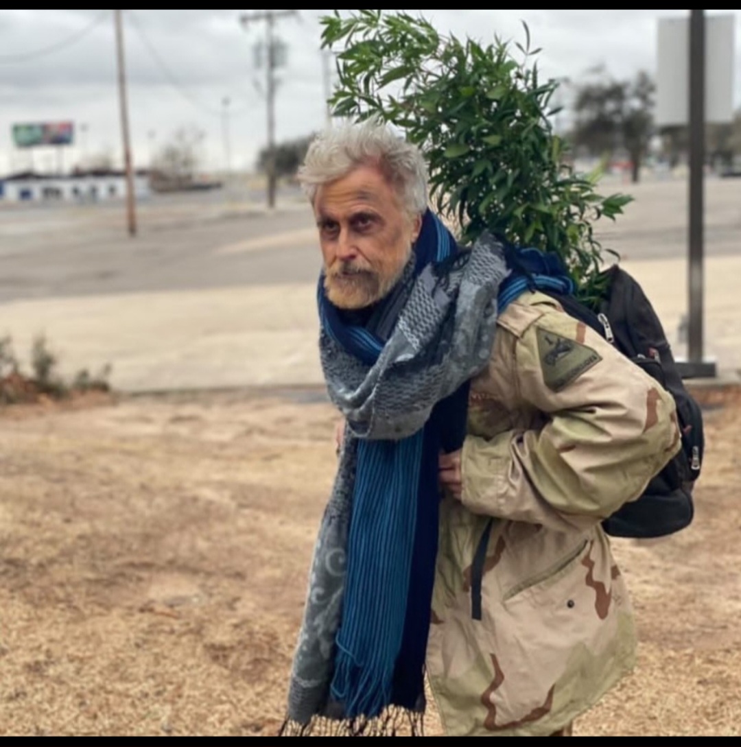 Older man with gray hair and beard, carrying a large plant on his back, wearing a beige jacket and scarf.