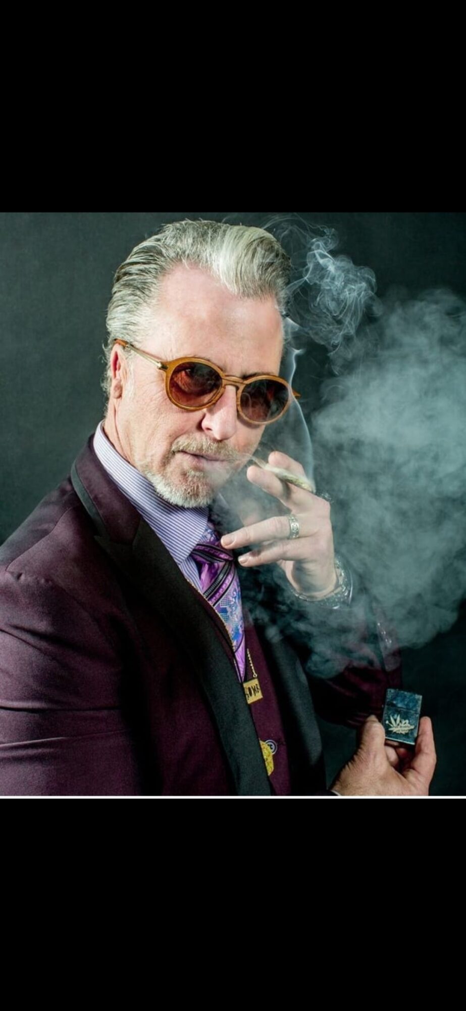 Man with sunglasses, smoking cigarette, wearing a suit and holding a lighter, with smoke around him.