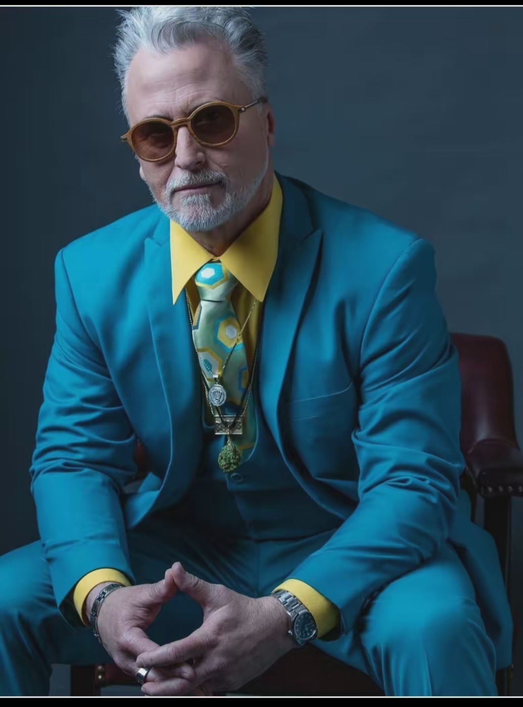 Older man with gray hair and beard wearing sunglasses, a bright blue suit, yellow shirt, and patterned tie, sitting with hands clasped.