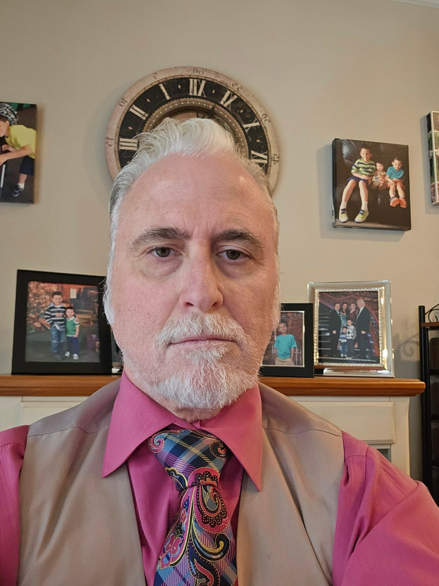 Man with gray hair and beard wearing a pink shirt and patterned tie, indoors with framed photos and a clock behind him.