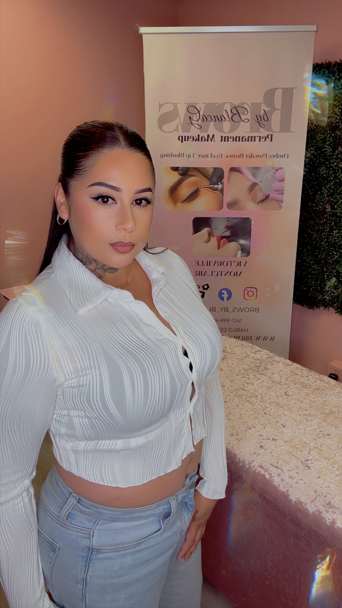 Woman with dark hair and makeup standing indoors near a promotional banner, wearing a white blouse and light blue jeans.