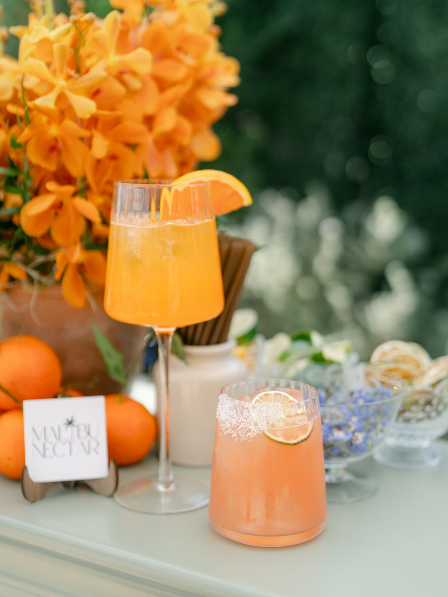 Two glasses with orange-colored drinks, one garnished with an orange slice, on a table with flowers and fruit.