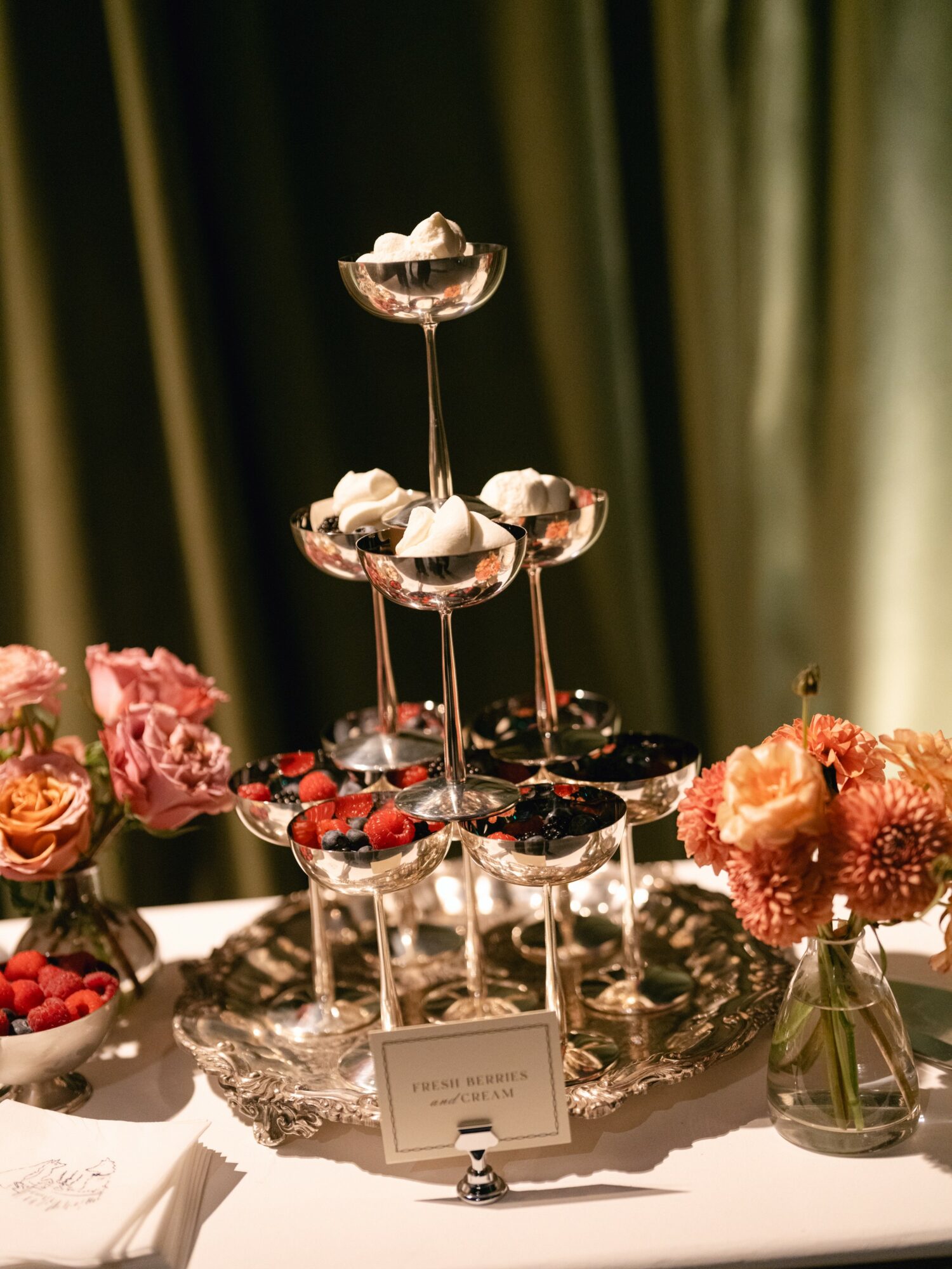 Elegant dessert display with glass stands holding chocolates and berries, surrounded by pink flowers in a vase, on a white table.