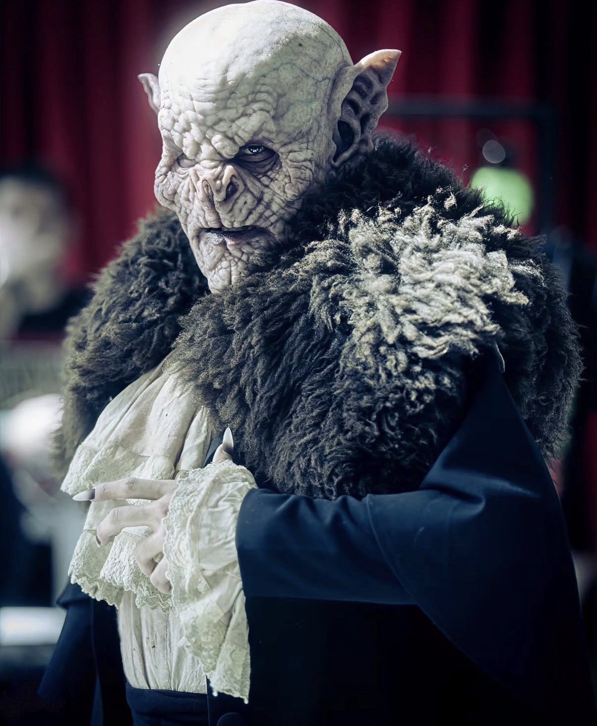 Person in detailed goblin costume with large ears, wrinkled face, and dark fur cloak, standing indoors with red curtains in background.