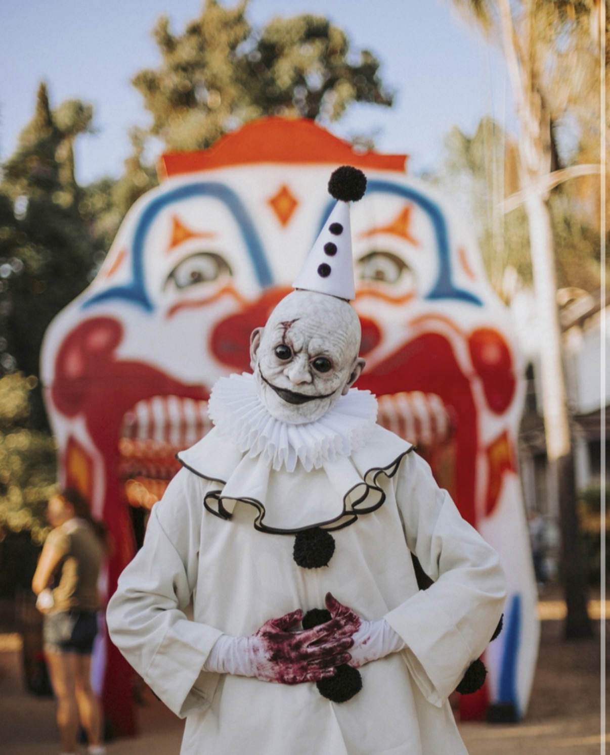 Person dressed as a clown with a painted face, wearing a white costume and a party hat, standing in front of a colorful carnival attraction.