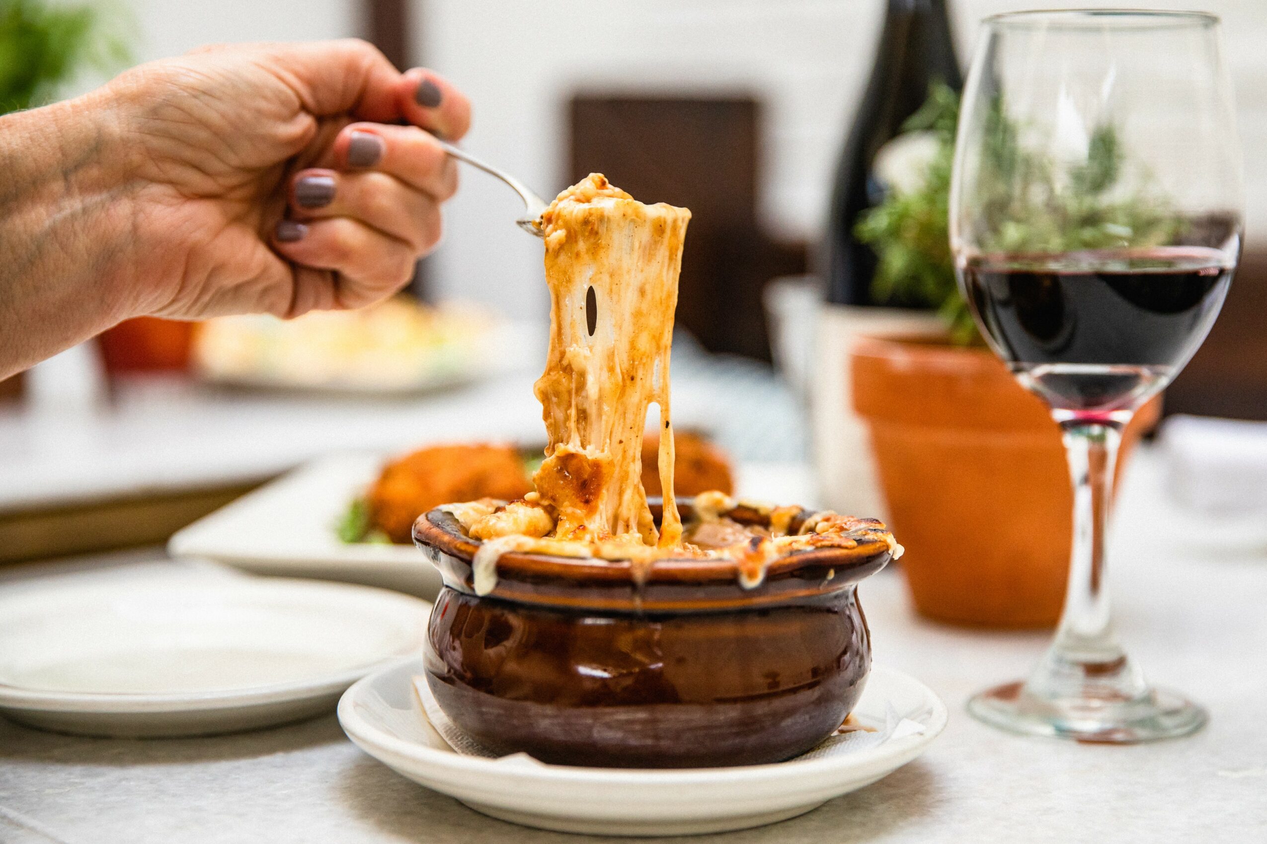 Person lifting a spoonful of cheesy, baked dish from a bowl on a table with a glass of red wine nearby.