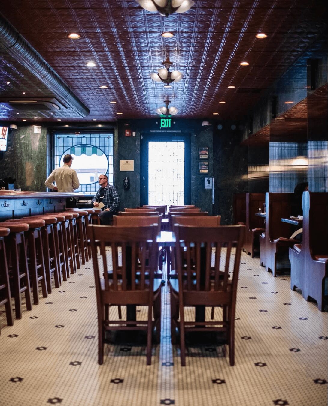 Empty dining area with wooden chairs and tables, bar counter on left, patterned floor, ceiling lights, and a door with an exit sign.