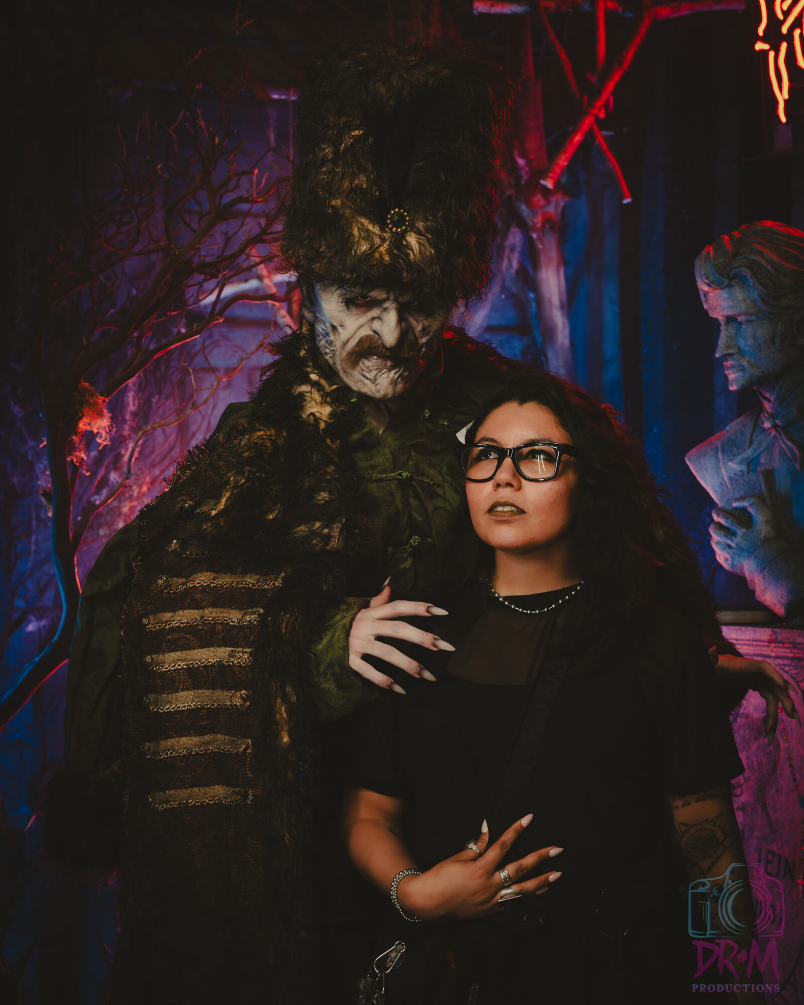 A woman with glasses and curly hair stands next to a person in a dark costume with makeup, in a colorful setting.