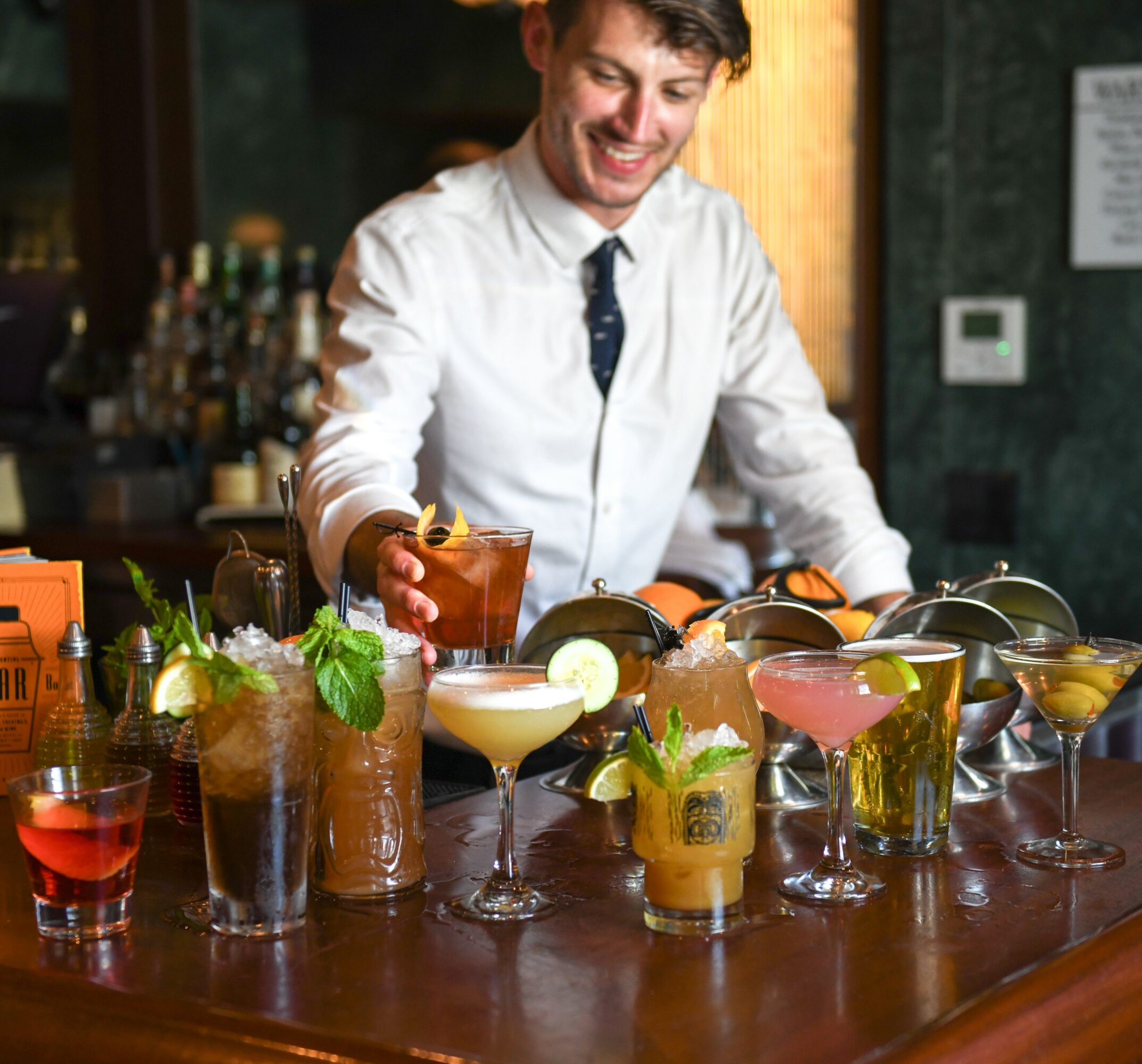 Bartender preparing colorful cocktails behind bar counter with various drinks and garnishes.