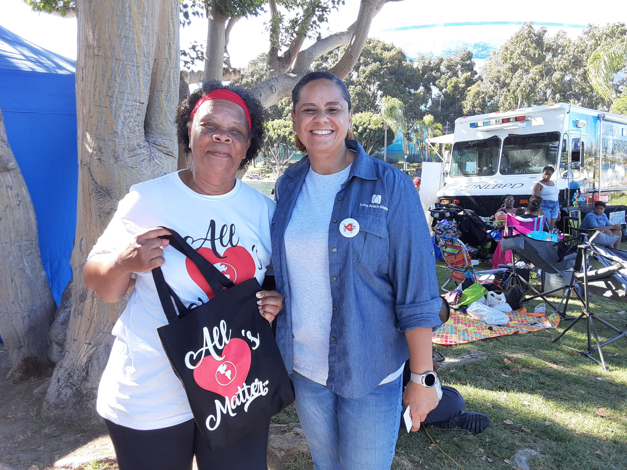 Two women smiling outdoors near trees and a bus, with chairs and items in the background.