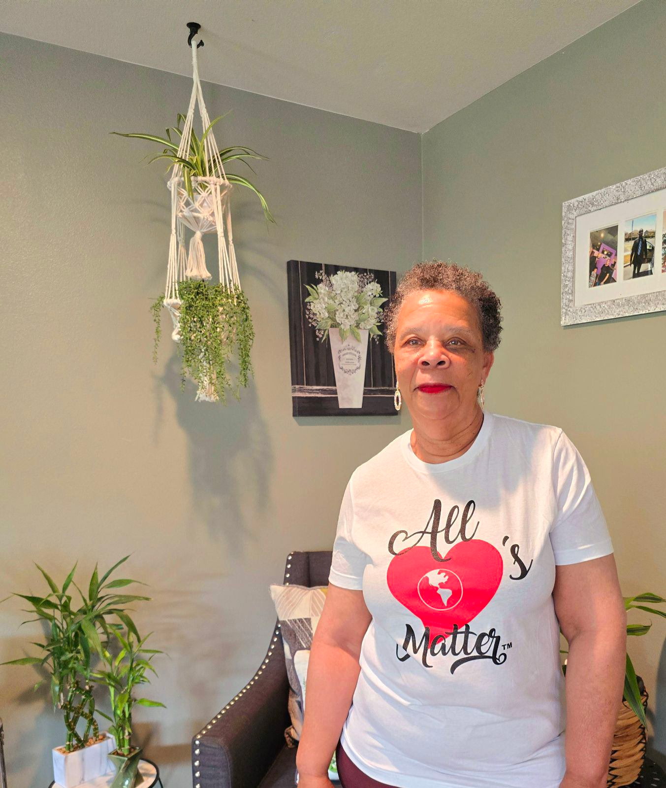 Older woman with short curly hair wearing a white T-shirt with red heart and text, standing indoors.