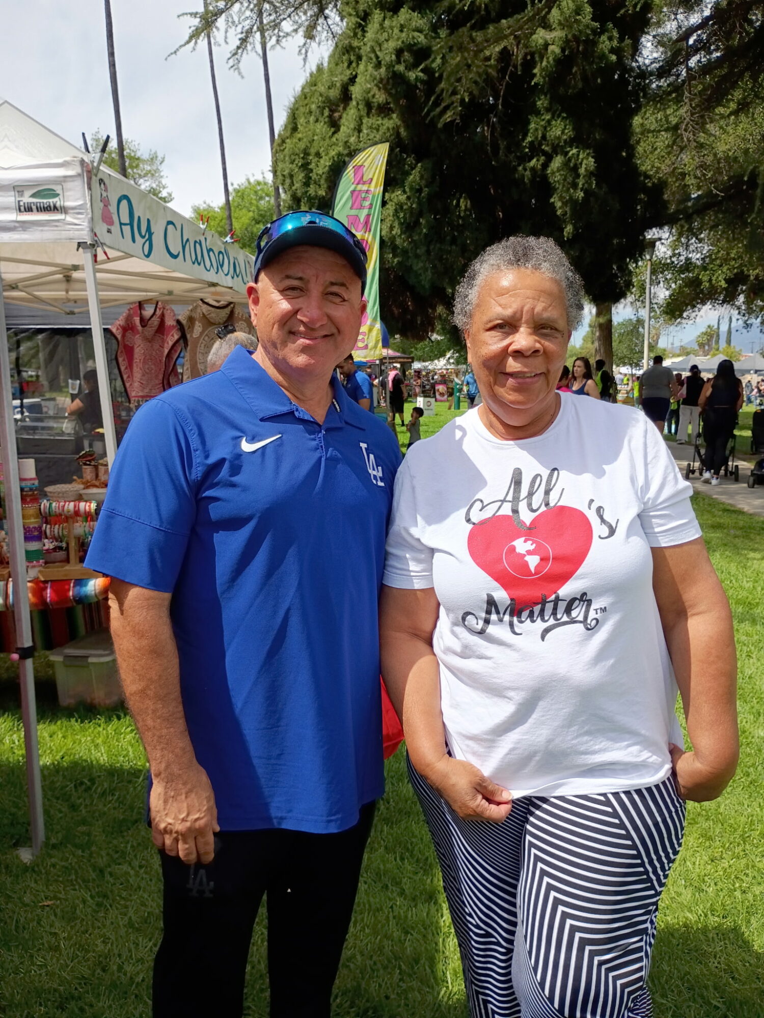 Two people standing outdoors, smiling, with tents and trees in the background.