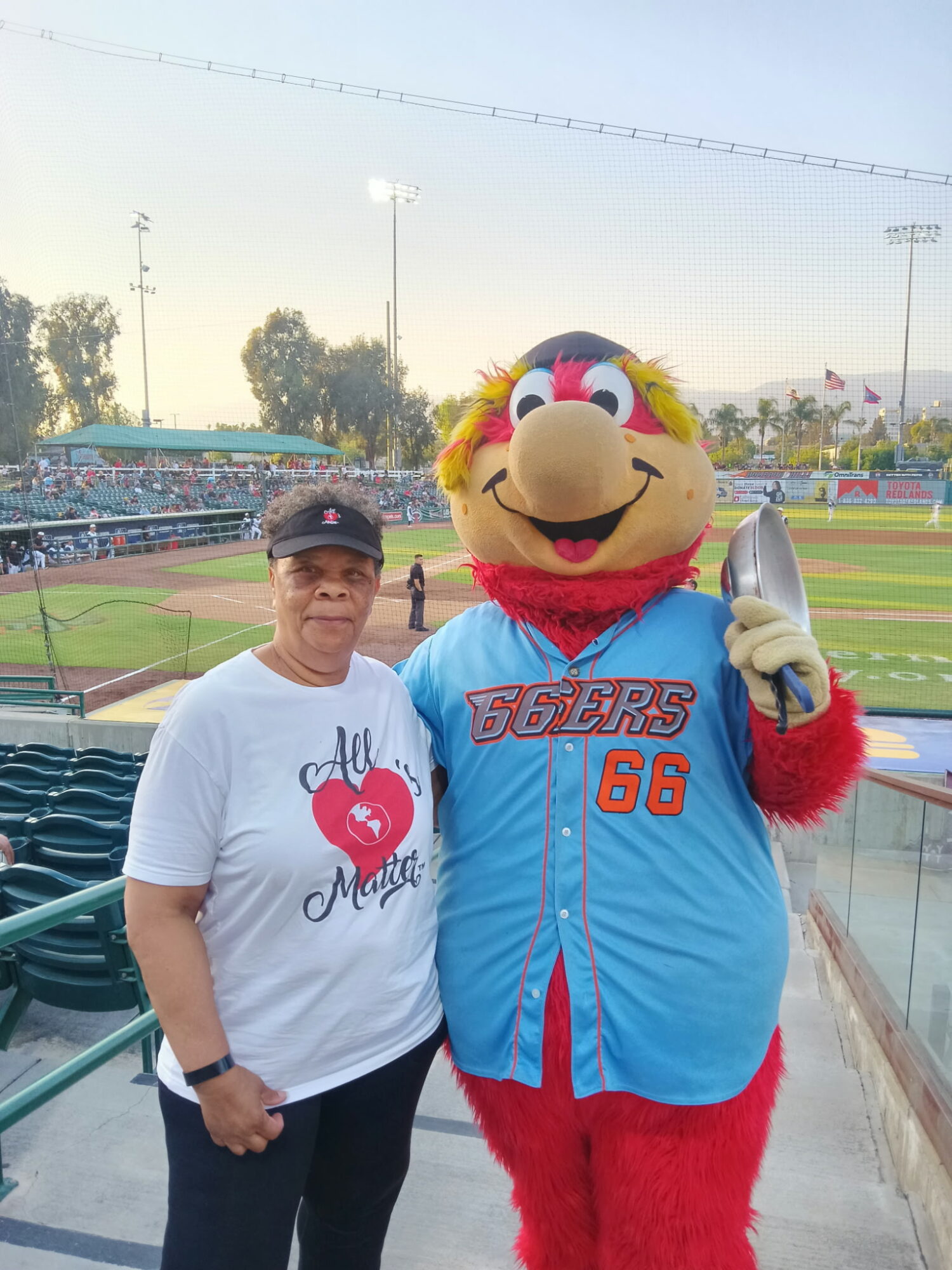 Person standing next to a colorful mascot in a baseball stadium, with empty seats and field in background.