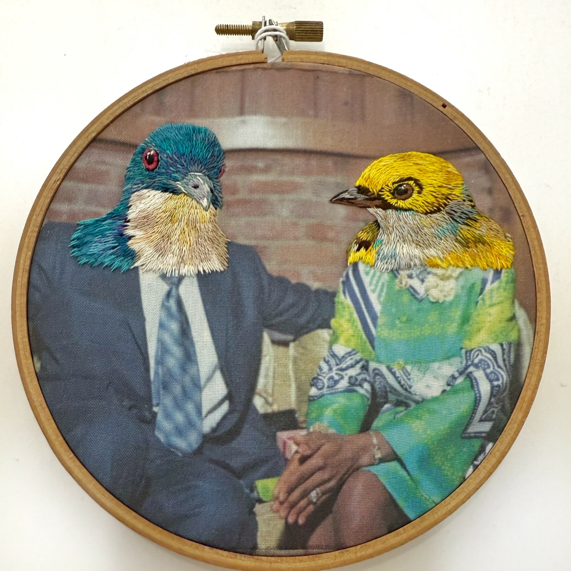 Two people with bird heads sitting together, one in a suit and the other in a colorful dress, against a brick wall.
