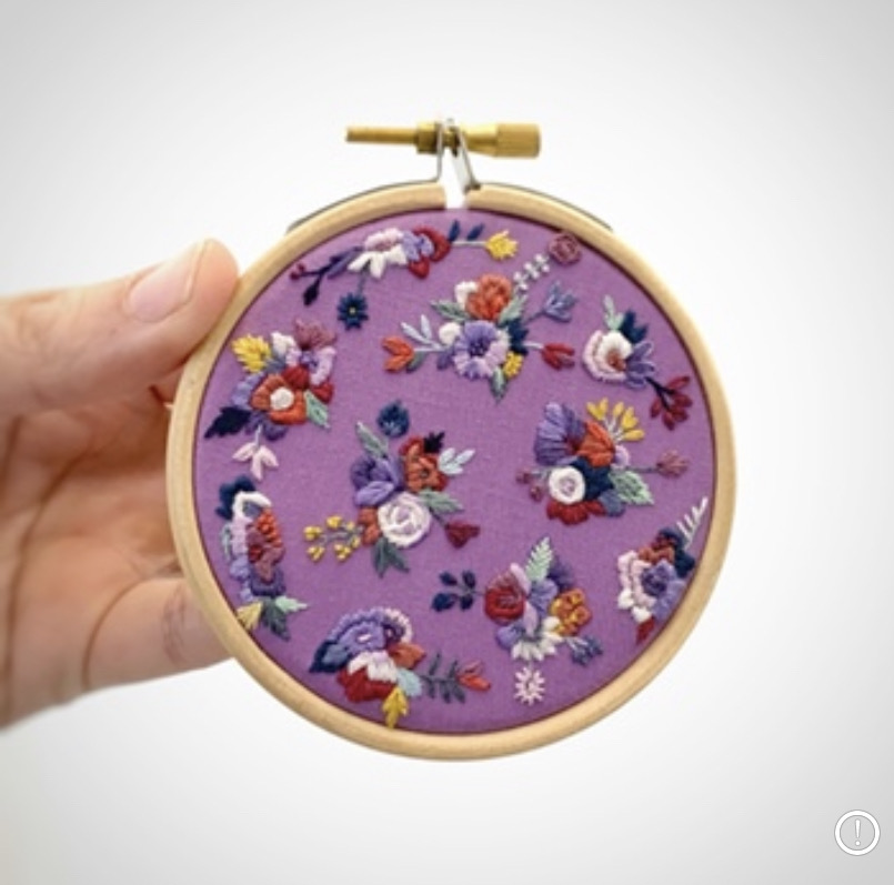 Hand holding a round embroidery hoop with purple fabric and colorful floral embroidery.