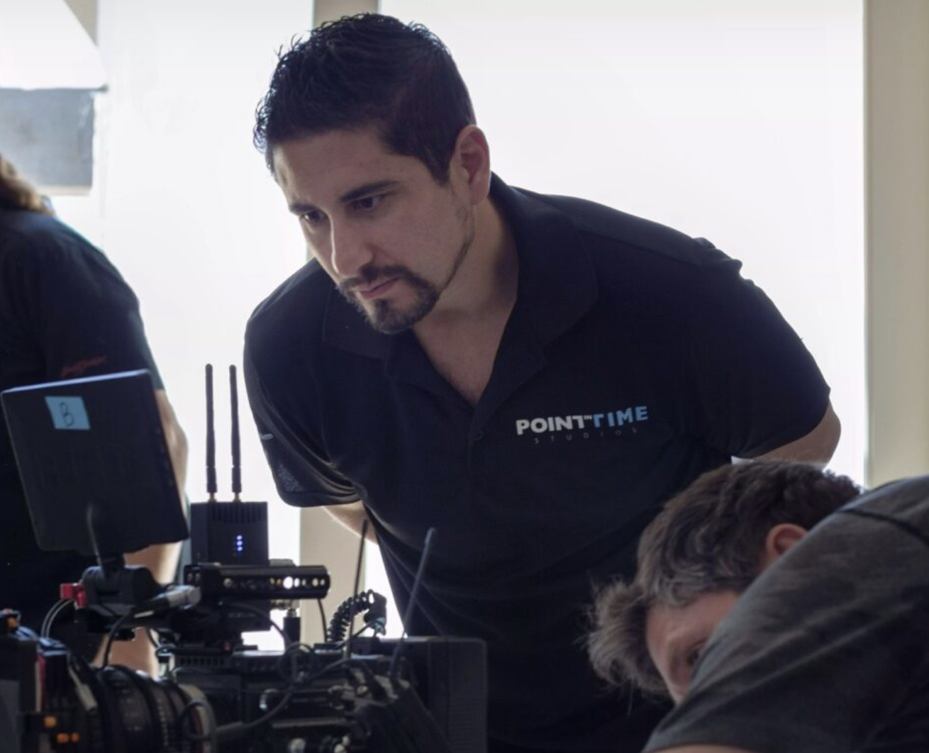 Man with dark hair and beard looking at camera equipment, wearing a black shirt with a logo.