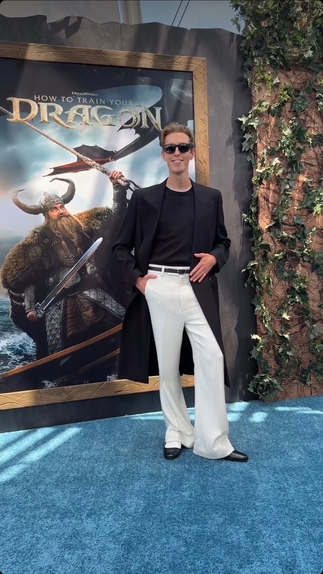 Person in black blazer, sunglasses, white pants, standing in front of a movie poster for 'How to Train Your Dragon'.
