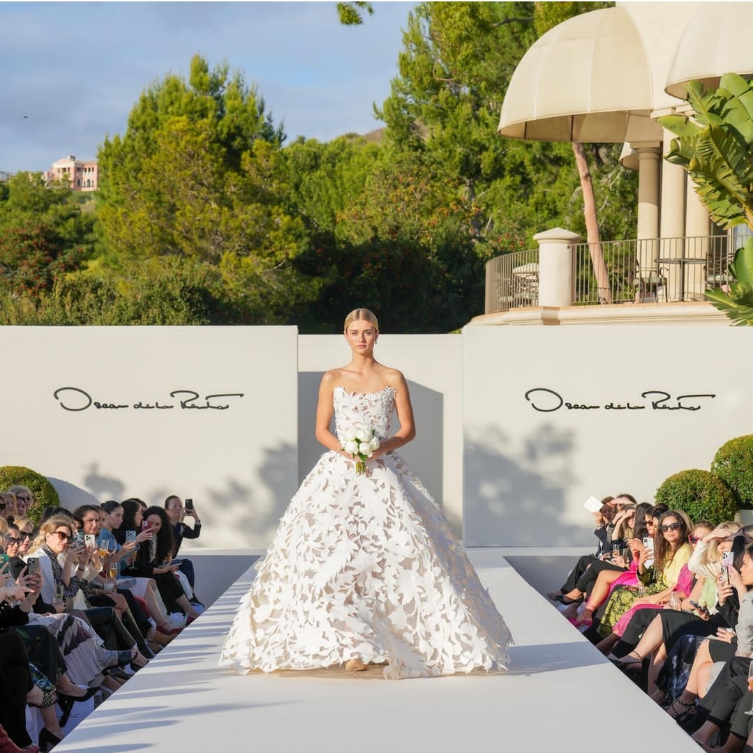 Bride in a white wedding gown holding a bouquet, standing on a runway outdoors with audience on both sides.