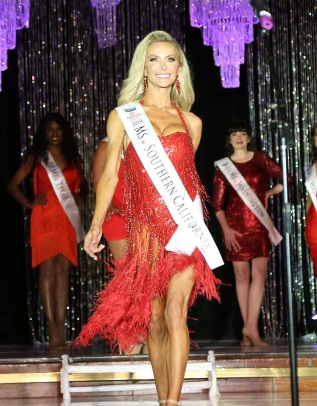 Woman wearing a sash and red dress on stage with two women in red dresses behind her, glittering backdrop.