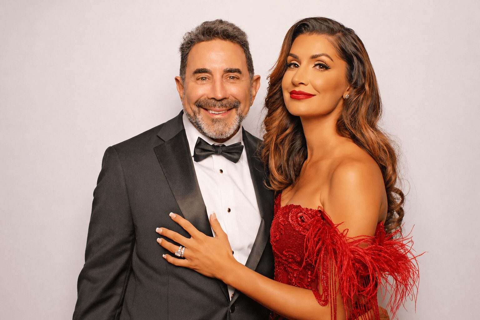 Man and woman in formal attire smiling, woman in red dress with feather details, man in tuxedo with bow tie.