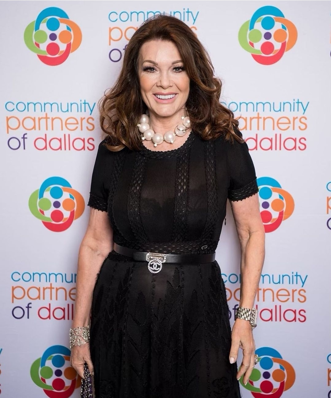 Woman with brown hair in a black dress and pearl necklace, smiling, standing in front of a colorful logo backdrop.