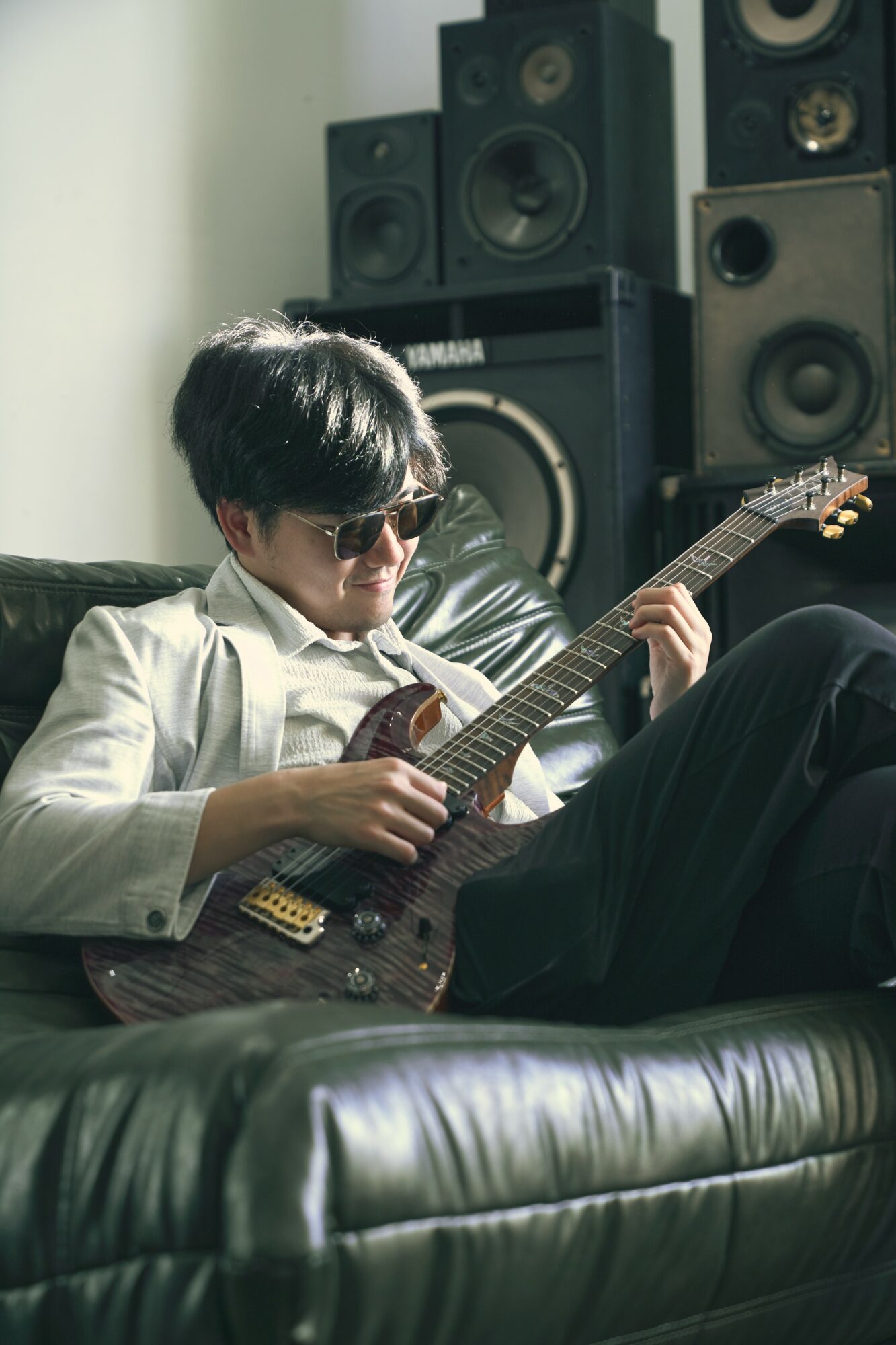 Person with dark hair and sunglasses playing an electric guitar on a black leather couch, surrounded by speakers.