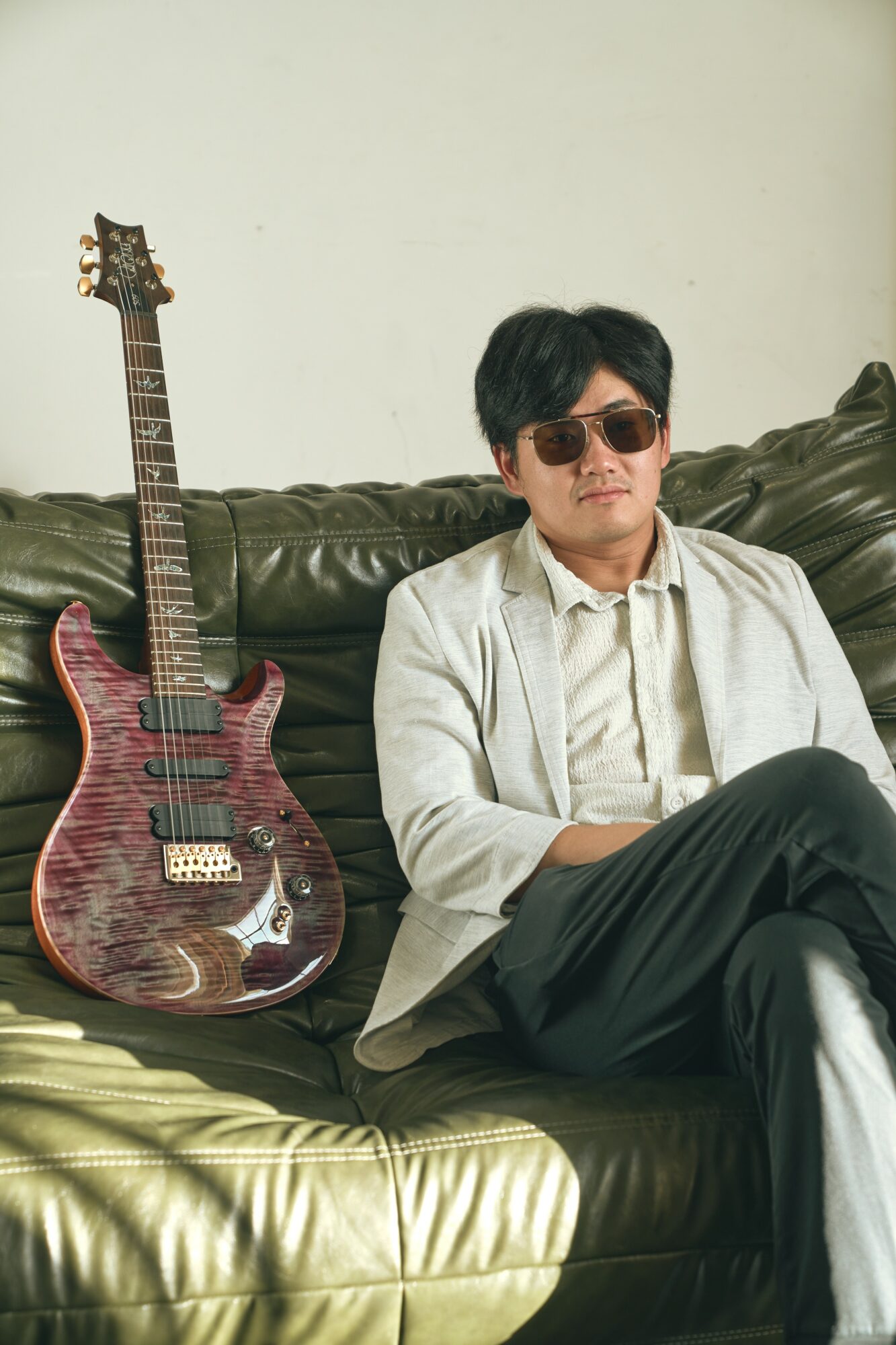 Man sitting on a green sofa with a guitar beside him, wearing sunglasses and a light-colored jacket.