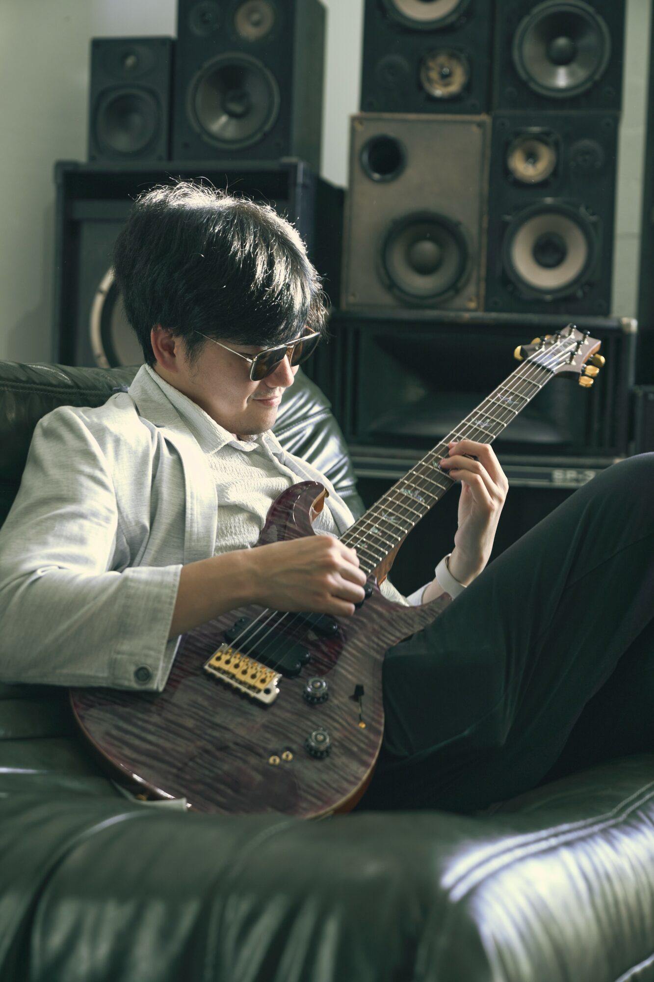 Person with dark hair and sunglasses playing an electric guitar in a music studio with large speakers behind.