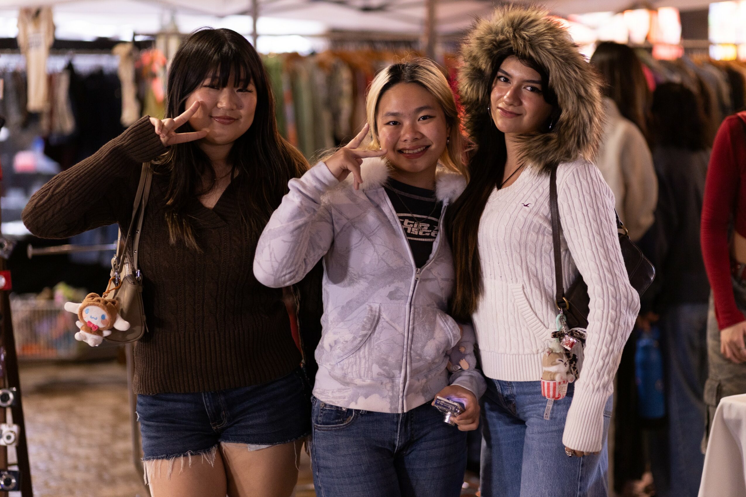 Three women smiling and posing at an outdoor market, with clothing stalls in the background.