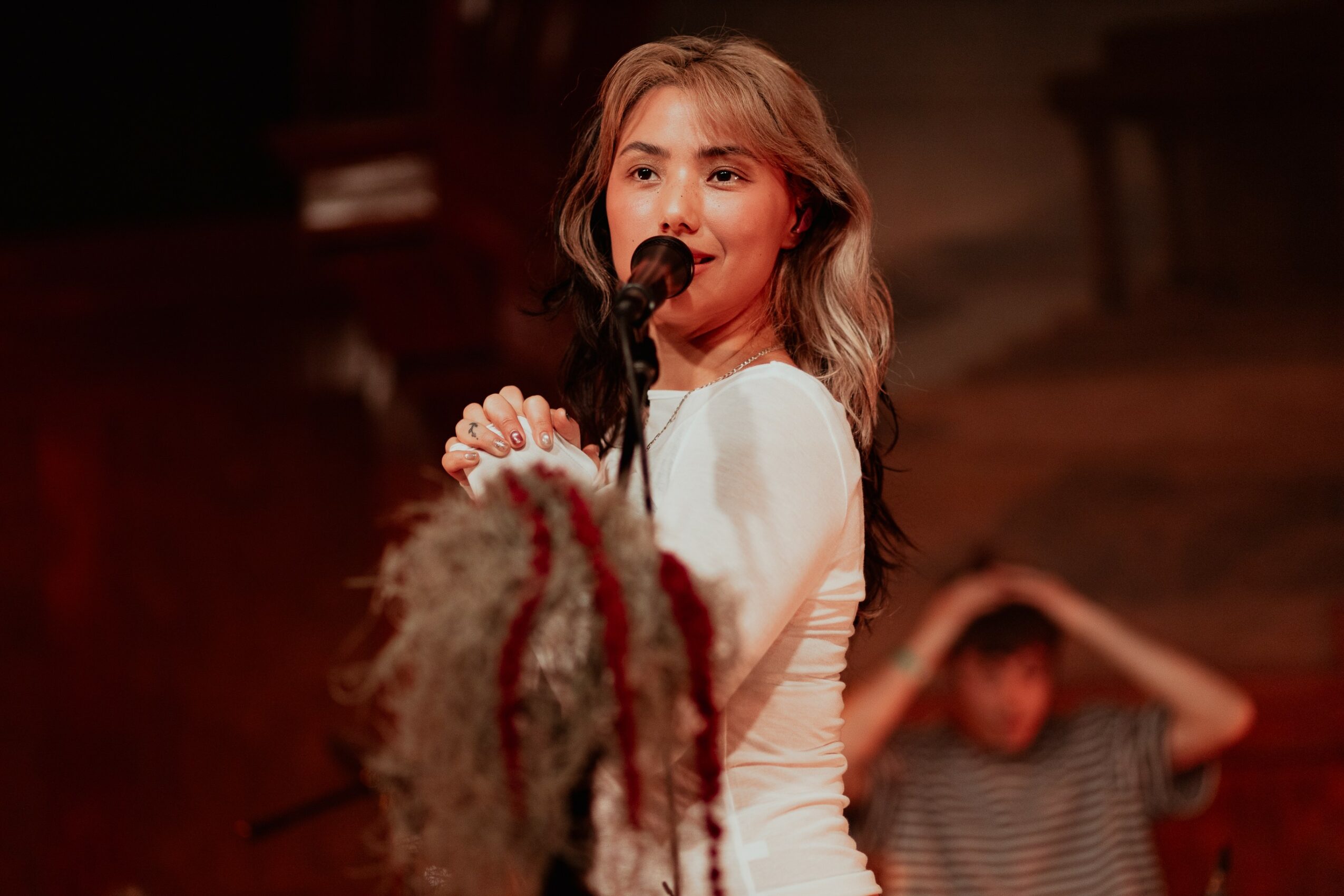 Young woman singing into a microphone on stage, with a person in the background holding their head.