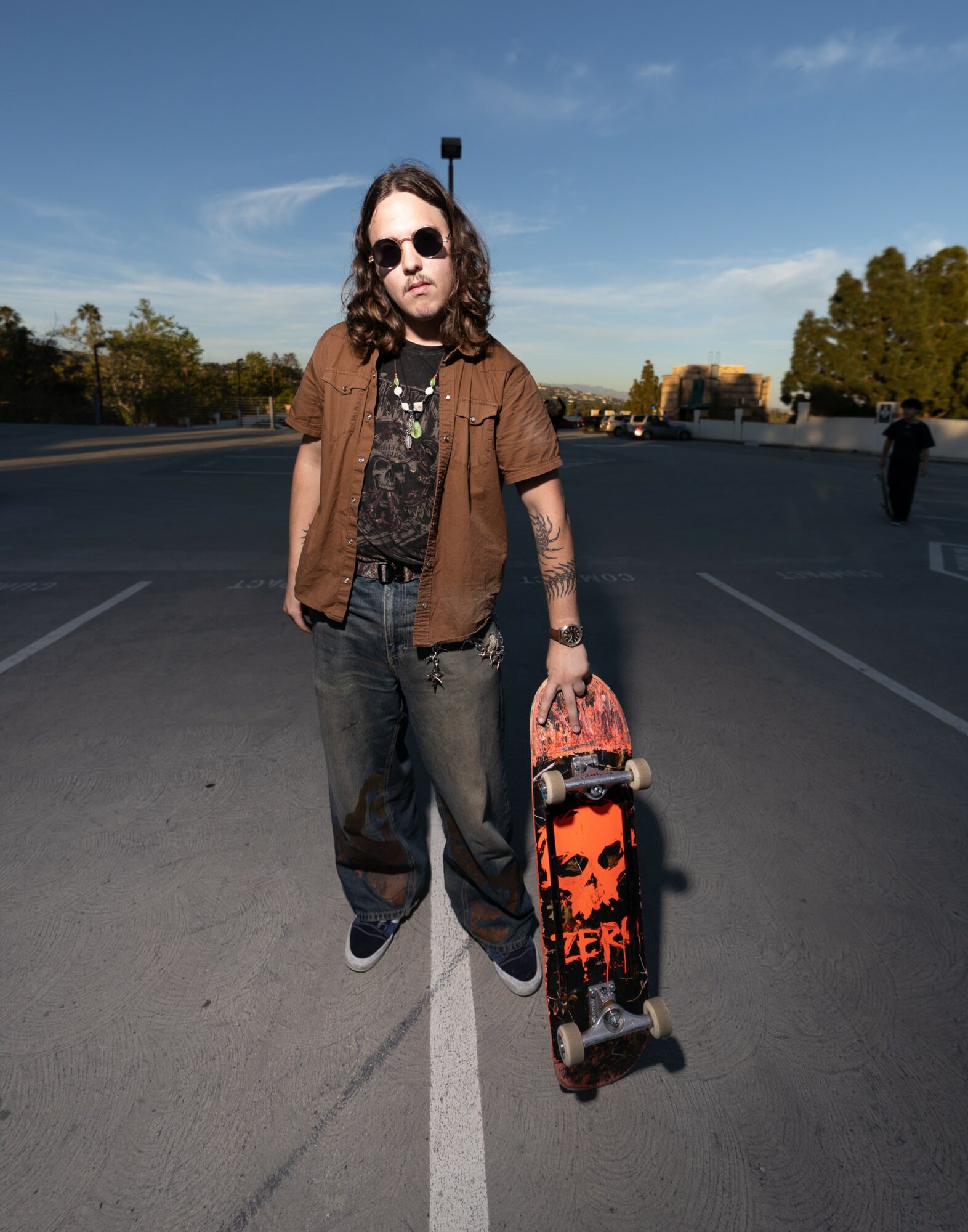 Person with long hair, sunglasses, brown shirt, and jeans holding a skateboard on a road.