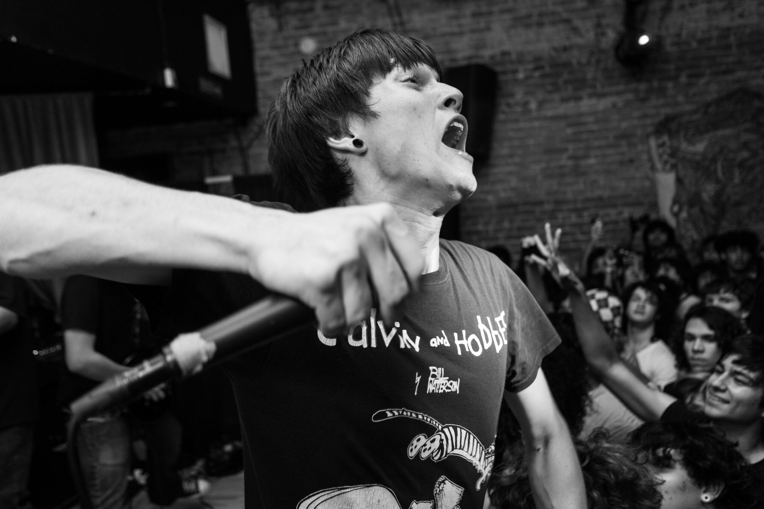 Person singing passionately with mouth open, holding microphone, crowd in background, black and white image.