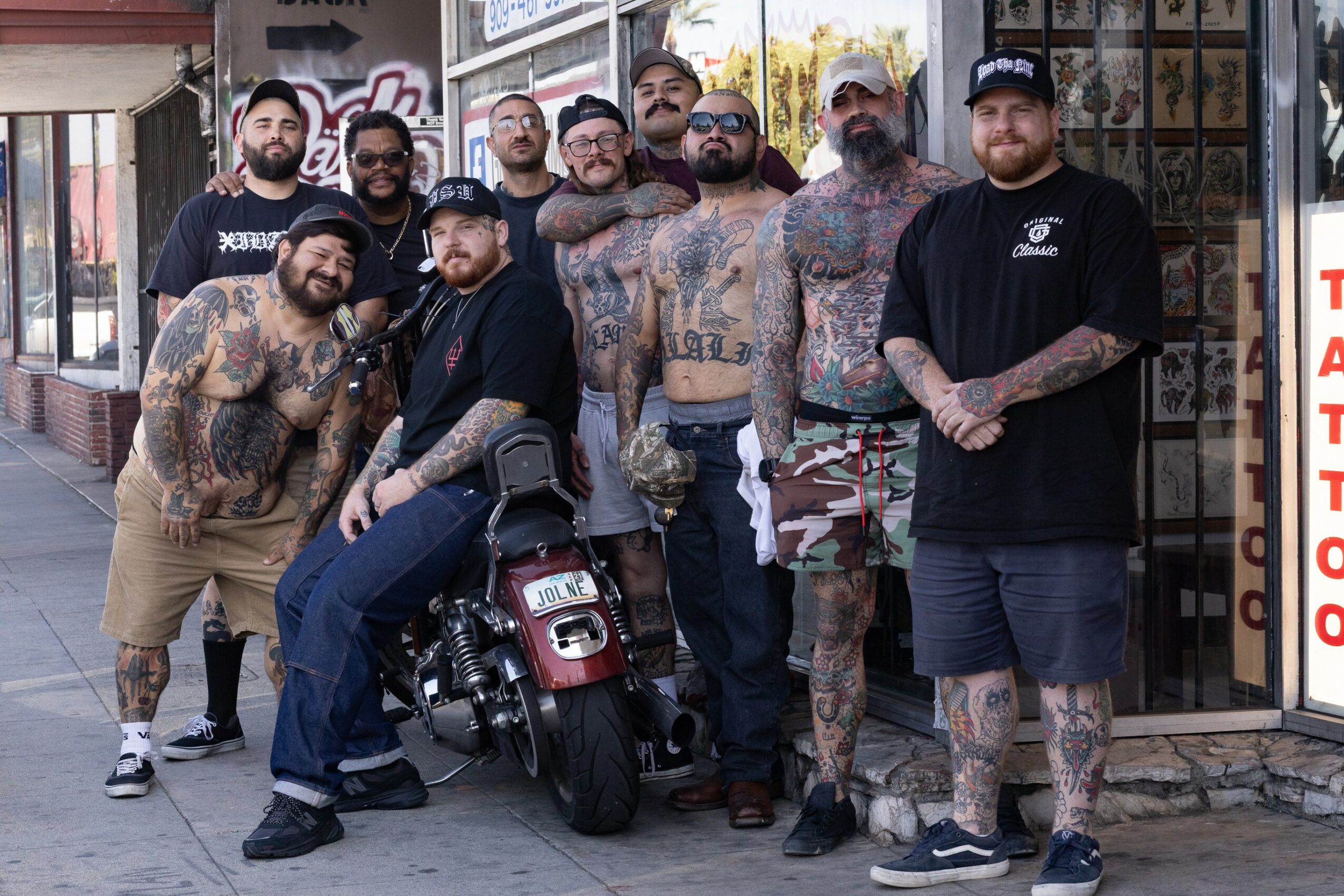 Group of ten men, some with tattoos, standing outside a building with glass windows and a door, some leaning on a motorcycle.