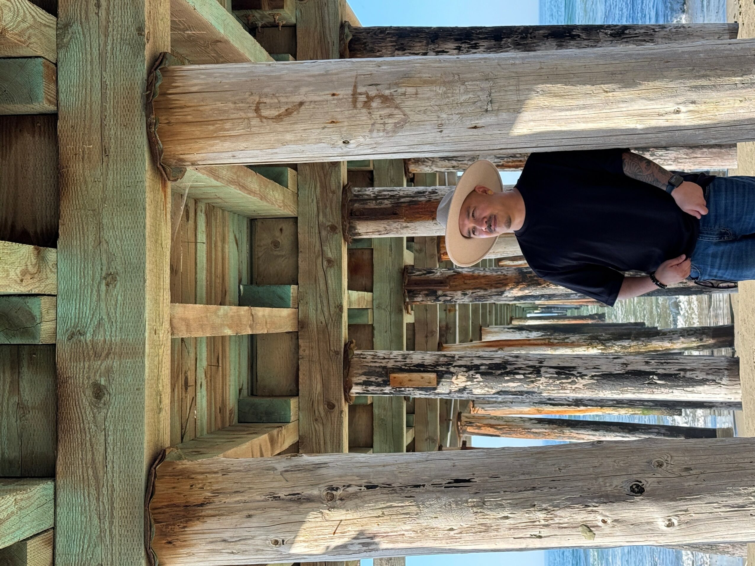 Person wearing a wide-brimmed hat and dark clothing standing among wooden posts and beams outdoors.