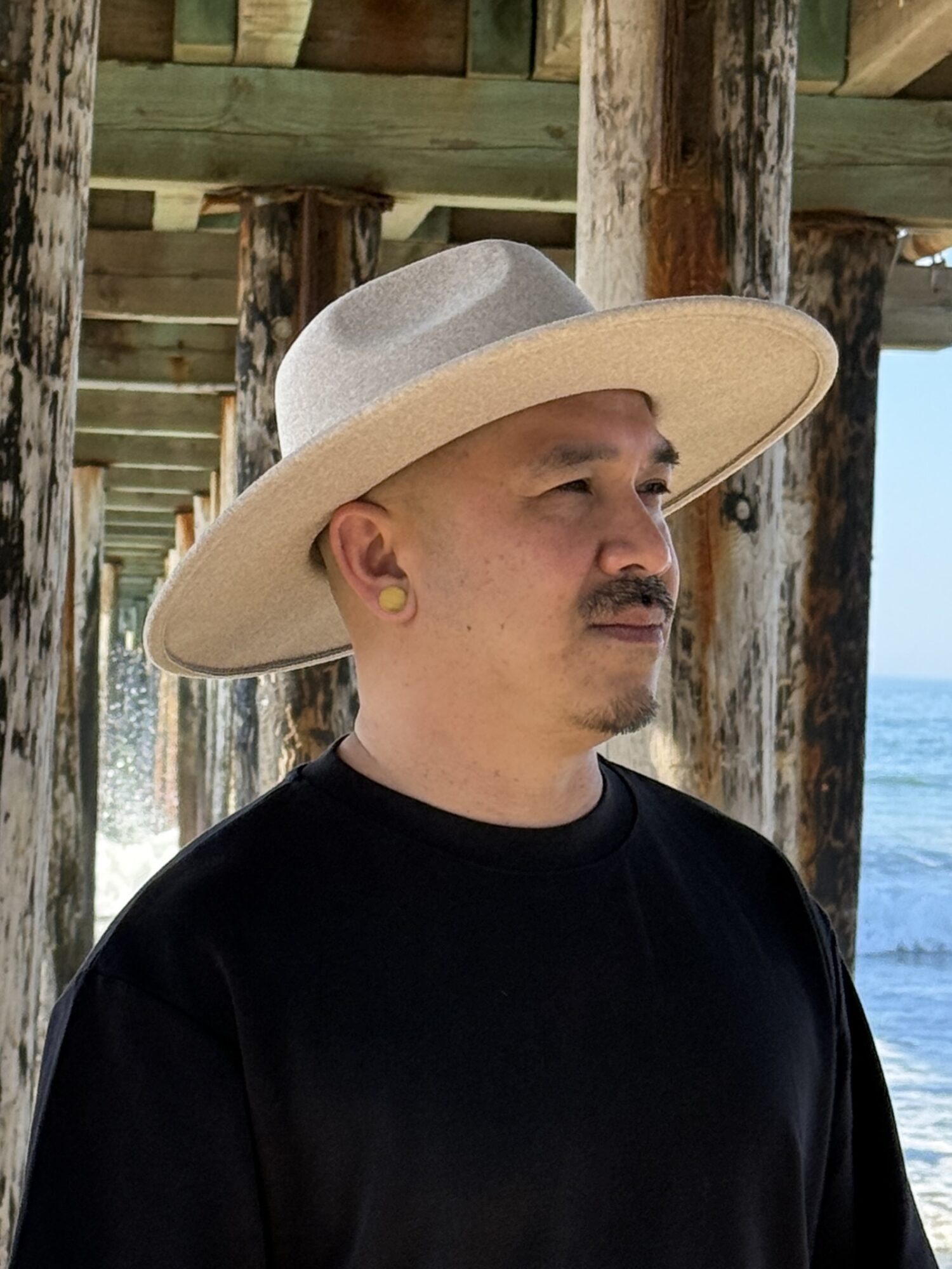 Man wearing a wide-brimmed hat and black shirt stands outdoors near wooden posts and water.