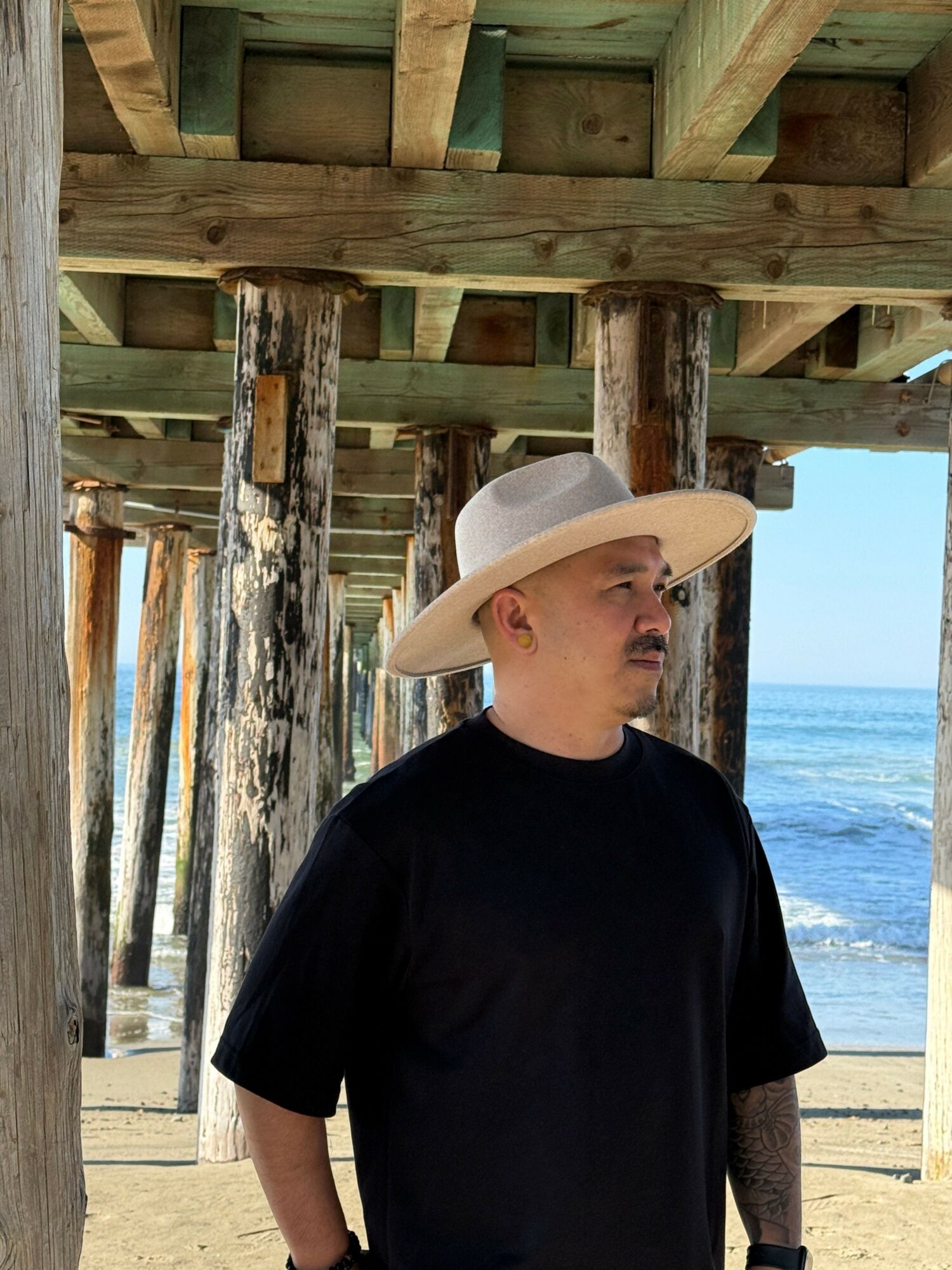 Man wearing a wide-brimmed hat and black shirt standing under a wooden pier near the ocean.