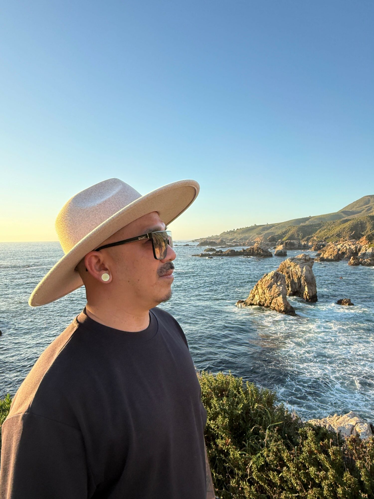 Person wearing a wide-brimmed hat and glasses stands near the rocky coastline during sunset.