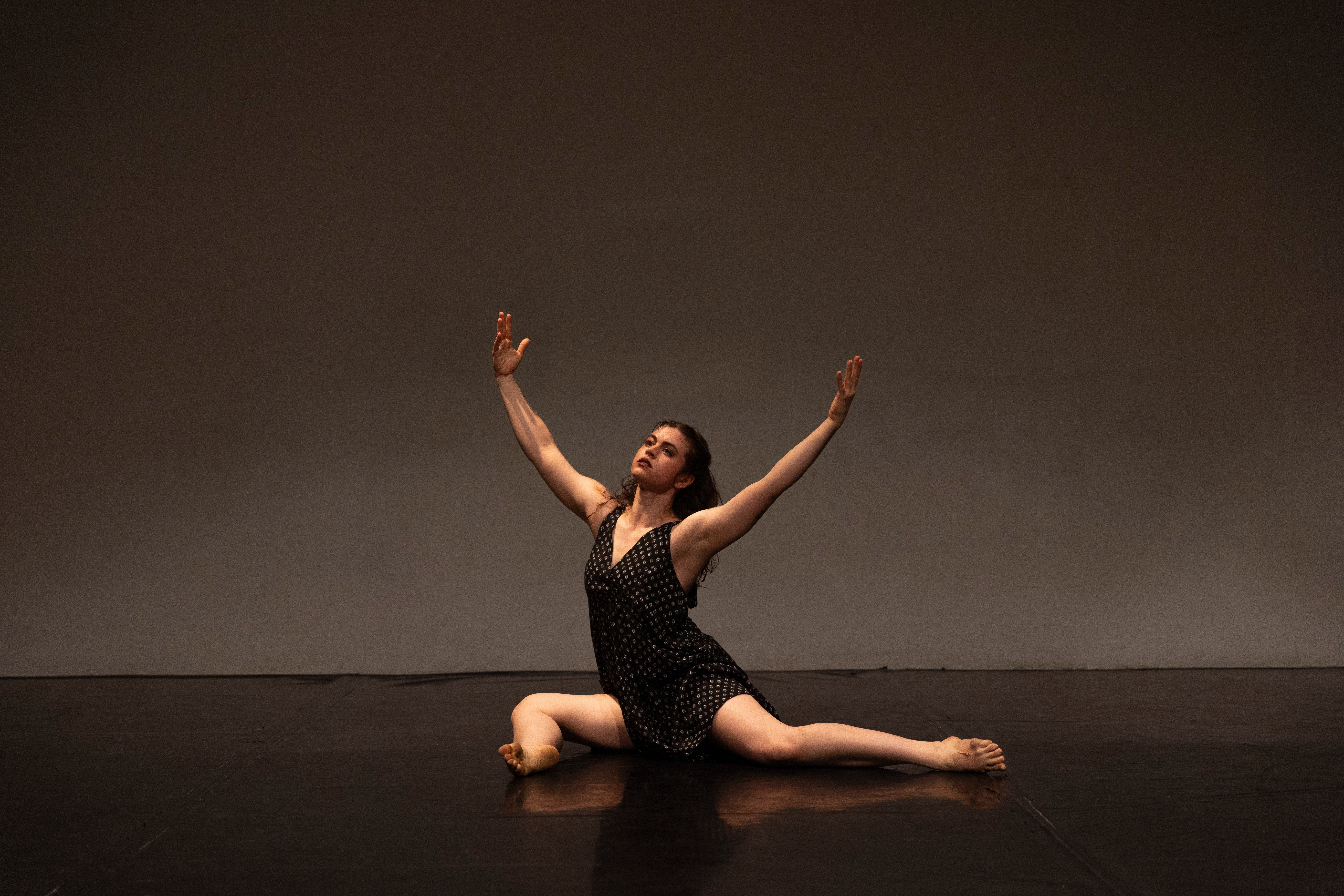 A woman performs a dance pose on stage, sitting with one leg extended and the other bent, arms raised.