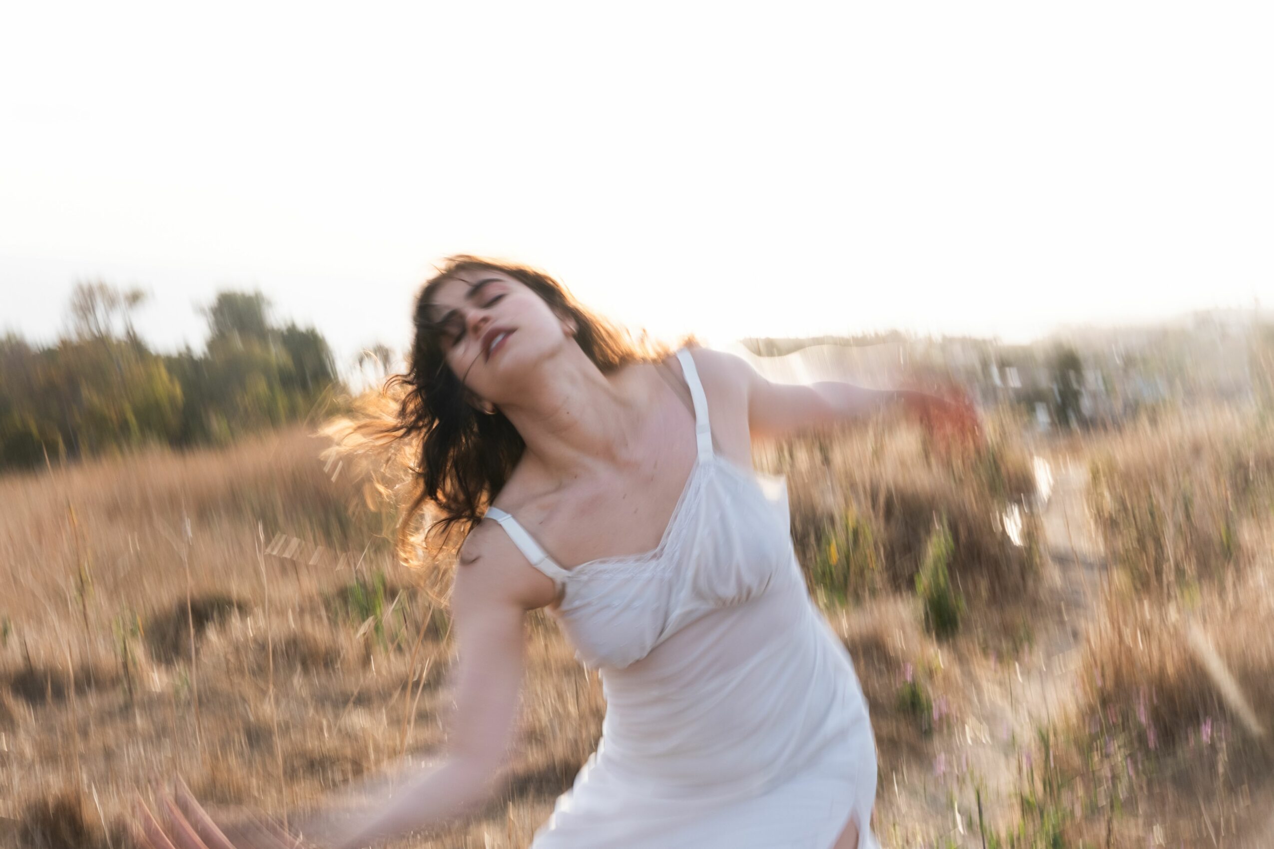 Woman in white dress with arms outstretched standing in a field during sunset.