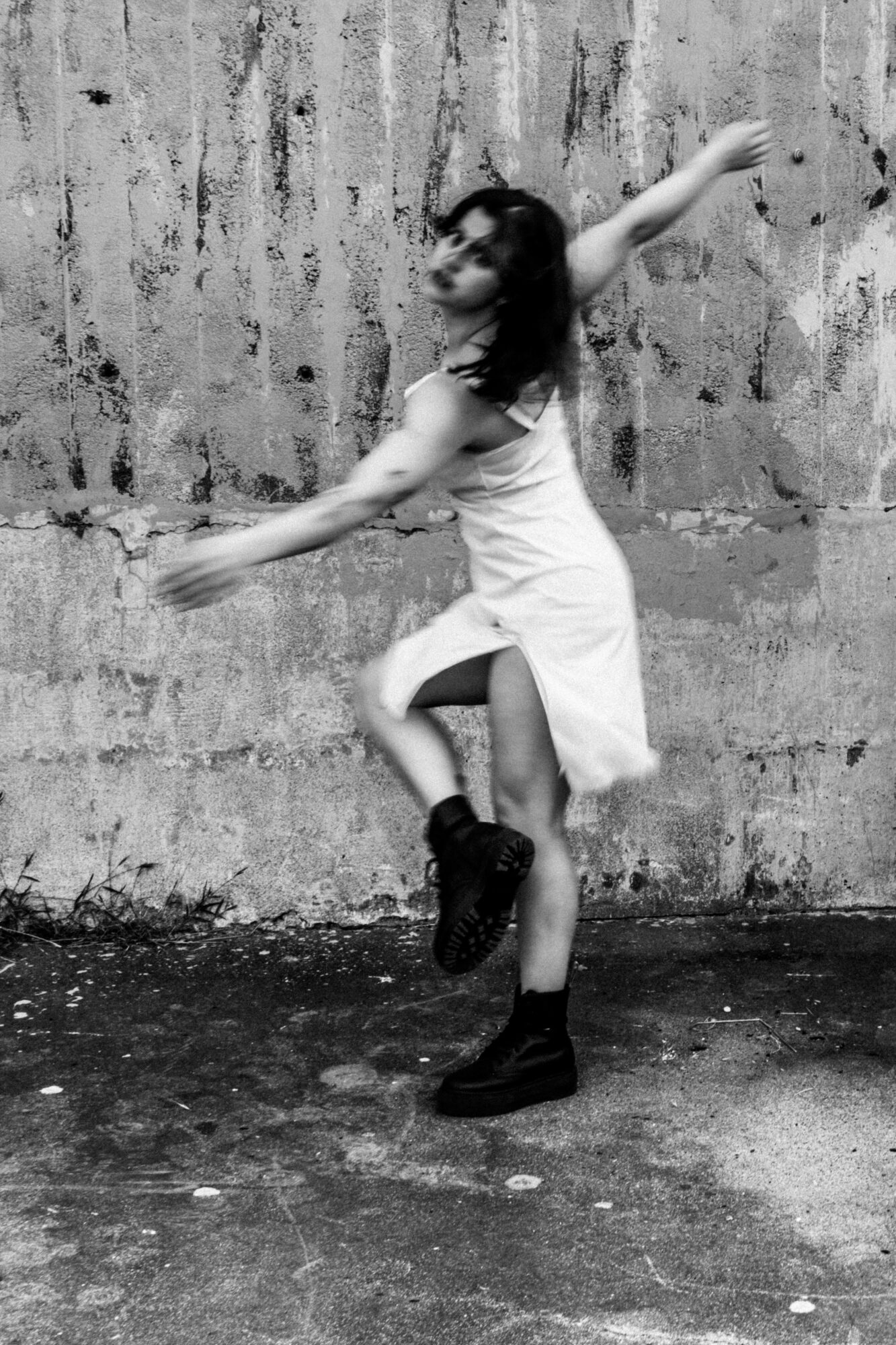 Young woman dancing outdoors against a textured wall, wearing a dress and boots, with arms and legs extended.