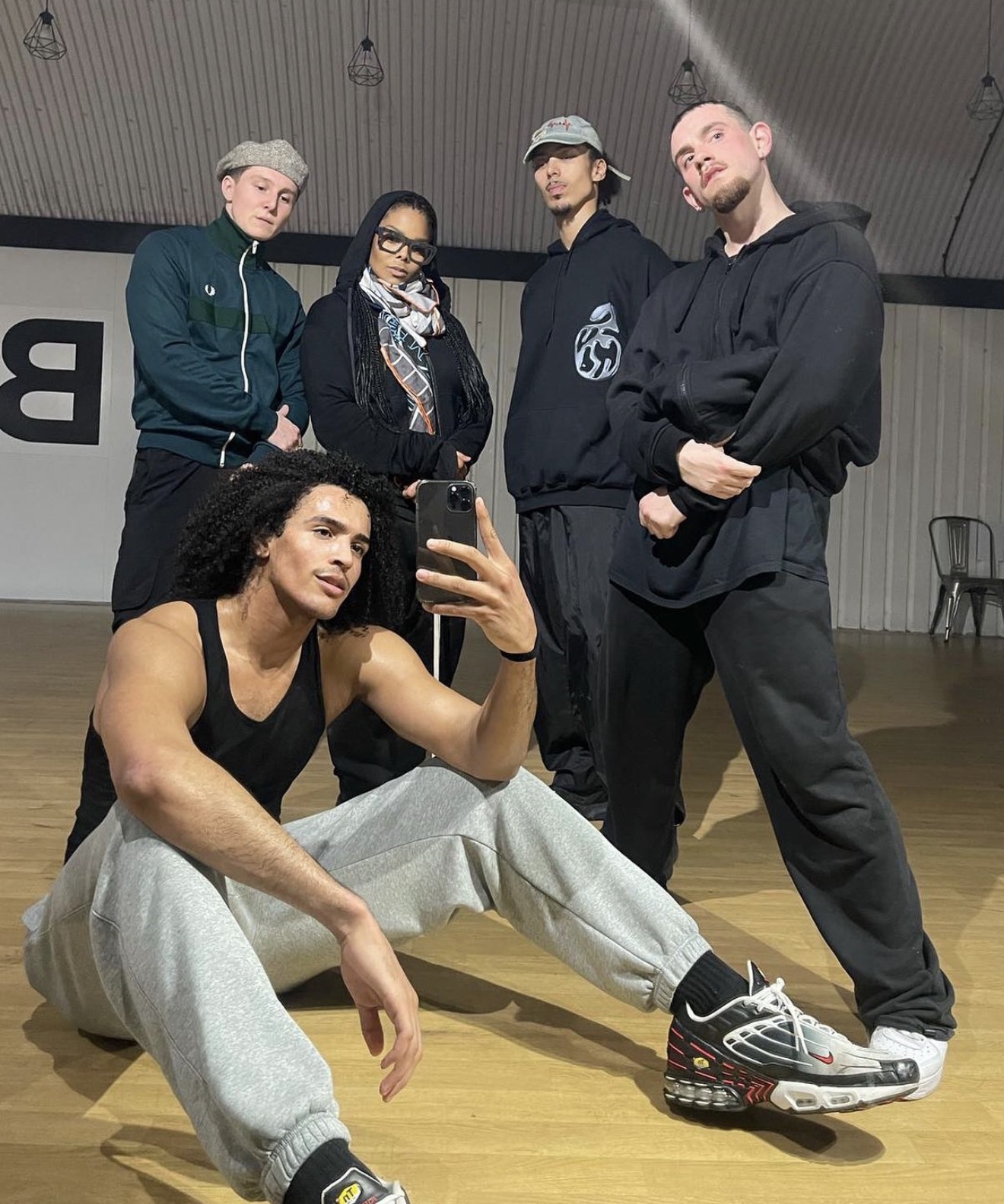 Five people in casual clothing posing indoors, one sitting on the floor taking a selfie, four standing behind.