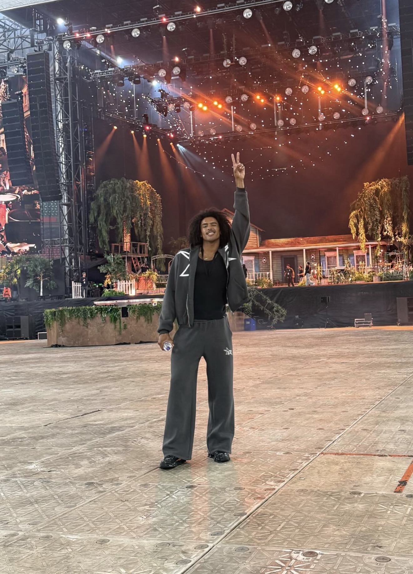 Person standing outdoors on stage with one arm raised, stage lights and trees in background.