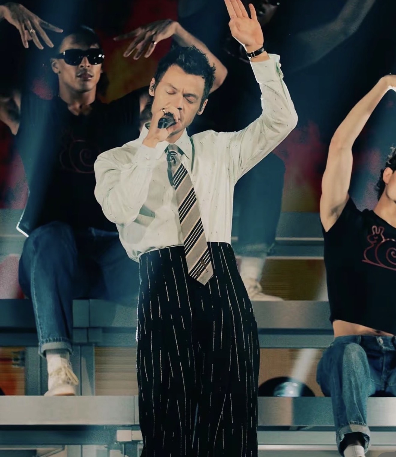 Man singing into microphone with raised hand, group performing on stage behind him, colorful background, casual attire.